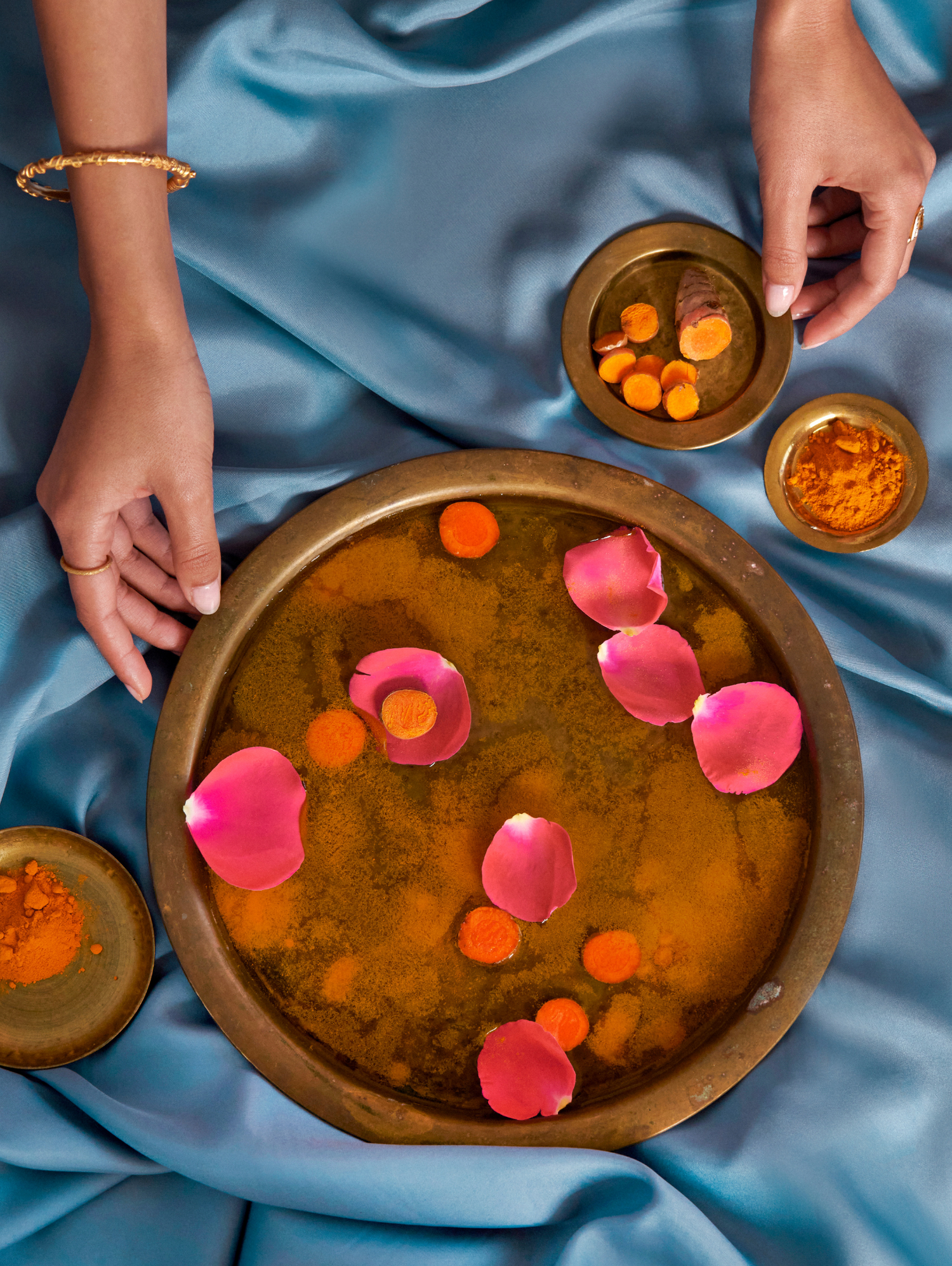 Hands hold and sift through turmeric root, turmeric powder, rose petals and water.
