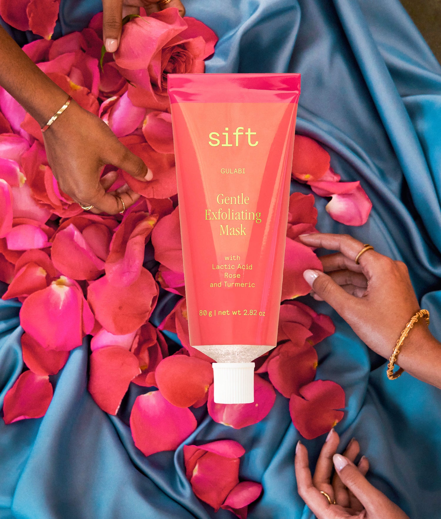 A tube of Sift's Gulabi, a gentle, exfoliating skincare mask, over hands holding rose petals. 